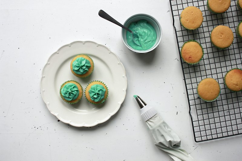 Drei mit Buttercreme dekorierte Cupcakes liegen auf dem Teller neben weiteren Cupcakes und eine Spritzbeutel. Mehrweg-Spritzbeutel sind oft aus Baumwolle oder Silikon und somit spülmaschinenfest.