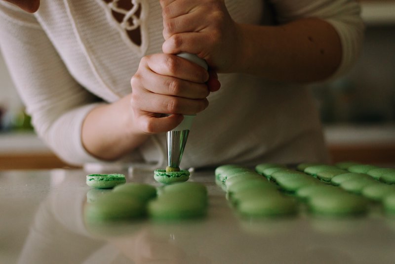 Frau befüllt grün gefärbte Macarons mit einer Creme mithilfe eines Spritzbeutels. Spritzbeutel kannst Du für Dekoration, Beschriftung oder für das Anrichten herzhafter Speisen verwenden.