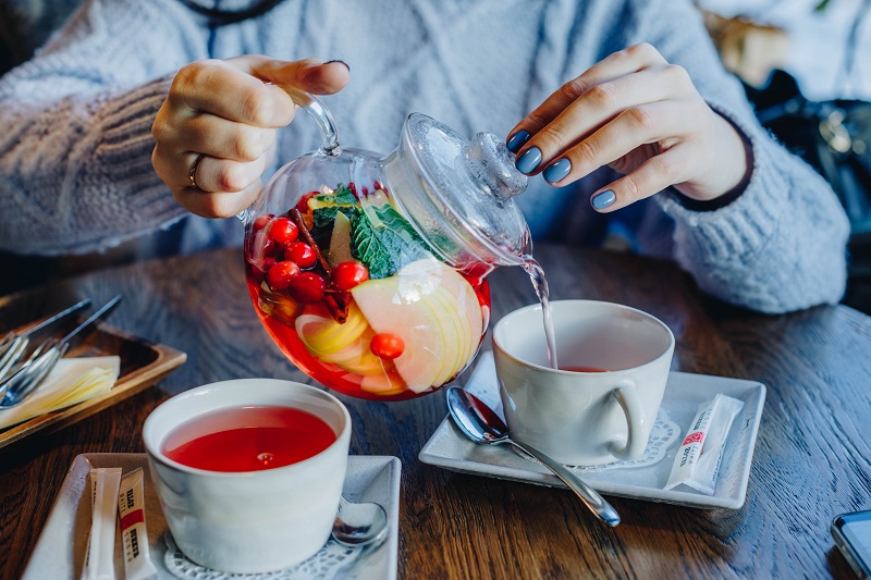 Frau gießt Tee aus Teekanne mit frischen Früchten in ihre Teetasse ein Teekanne mit frischen Früchten