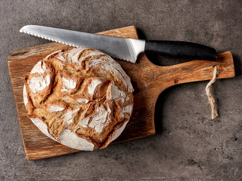 Brötchenmesser neben einem frischen Brotlaib Frischer Brotlaib auf einem Brett neben einem großen Brotmesser