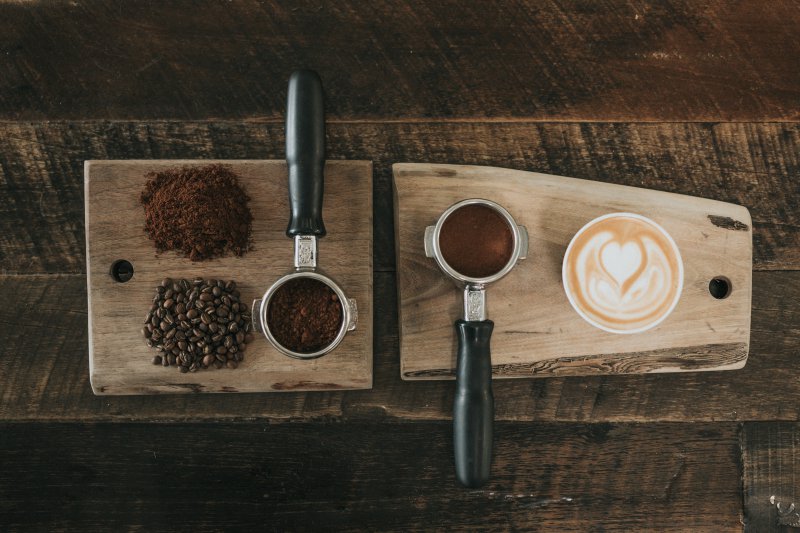 Cappuccino, Kaffeebohnen und Espressopulver in Siebteilen auf einem Holzbrett Cappuccino und Kaffeebohnen/pulver