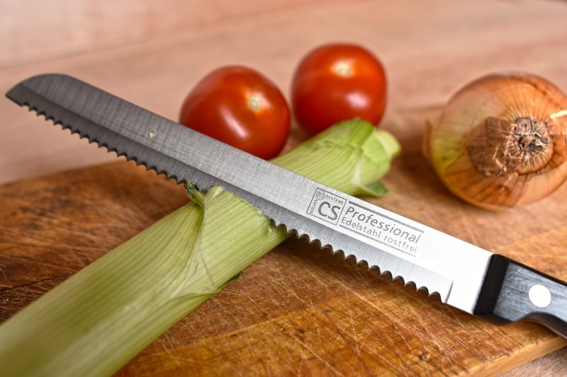 Großes Tomatenmesser mit einer Zwiebel Großes Tomatenmesser schneidet in Lauch neben einer Zwiebel und Tomaten auf einem Holzschneidebrett